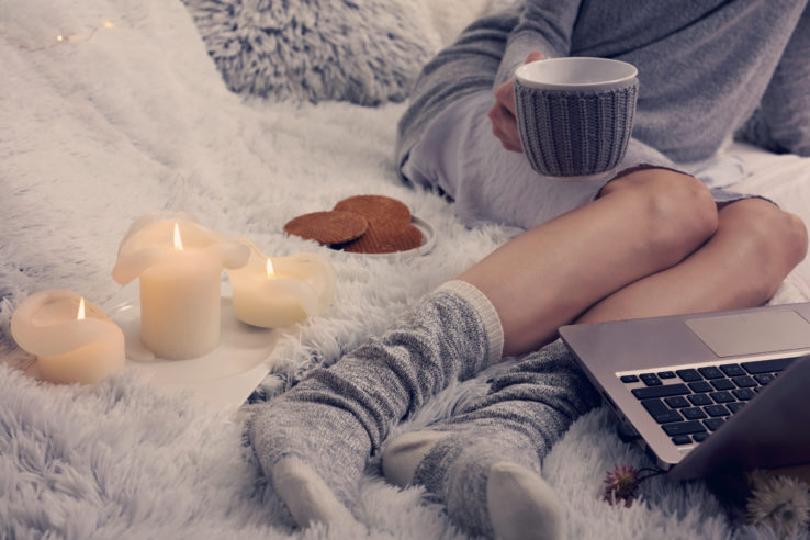 Cozy evening , warm woolen socks, soft blanket, candles. Woman relaxing at home,drinking cacao, using laptop. Comfy lifestyle.